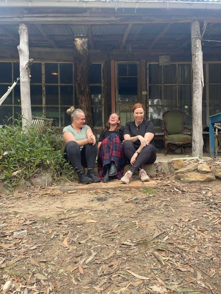 Three ladies in their 40s sitting in front of an old wood cabin at Darlinghurst Farm having a laugh